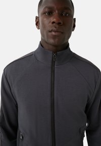 Ciemnoszara kurtka zapinana na zamek z wysokim kołnierzem, teksturowana tkanina, ukośne detale szwów i kieszenie boczne na zamek. Charakteryzuje się smukłym designem.