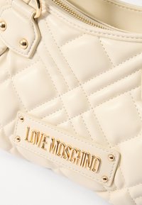 Quiltana kremna torba iz umetnega usnja z zlatimi dodatki, ki vsebuje oznako "LOVE MOSCHINO" in zadrgo na vrhu.
