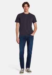 T-shirt di cotone blu scuro a maniche corte, abbinata a jeans in denim medio e sneakers nere e bianche, in piedi su uno sfondo semplice.