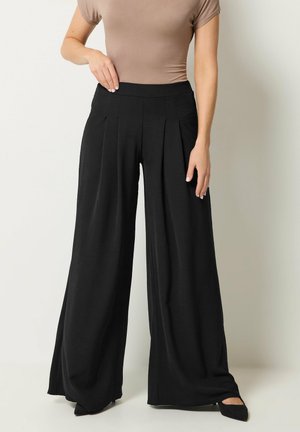Woman wearing high-waist wide-leg black pants with pleats, paired with a beige fitted short-sleeve top and black pointed shoes.