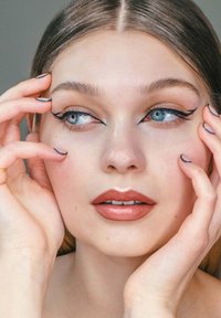 Close-up van een gezicht met lichte huid, blauwe ogen en duidelijke zwarte eyeliner. Lippen zijn een zachte roze tint met een glanzende afwerking. Handen omlijsten het gezicht.