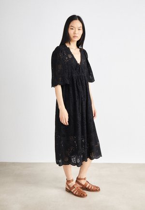 Black cotton dress featuring a v-neck, puff sleeves, eyelet cut-out patterns, and a flared hem; paired with brown strappy sandals.