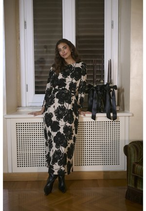 Fekete és bézs virágmintás ruhát viselő nő fekete csizmában, aki egy ablakpárkányra támaszkodik, mellette három magas fekete gyertya szalaggal megkötve.