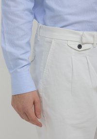 Torso maschile con camicia a maniche lunghe blu chiaro e pantaloni bianchi a vita alta con un bottone nero e tasche laterali.