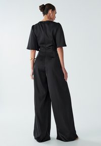 Sieviete ar matiem kopā un melnā platajā kombinezonā ar elkoņa garuma piedurknēm, zeltainiem rokassprādzēm un auskaru pāri, stāv uz balta fona.