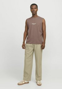 Camiseta sin mangas marrón con el texto blanco "Originals Studio", combinada con pantalones de pierna ancha beige y zapatillas beige con acentos claros.