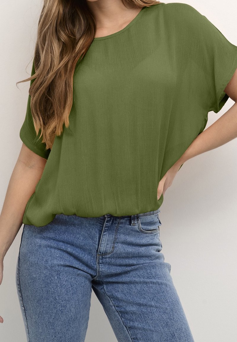 Haut olive, à manches courtes et coupe ample, en tissu texturé. Associé à un jean bleu à taille haute avec des poches sur le devant.