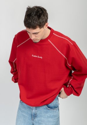 Giovane uomo con capelli corti e scuri che indossa una felpa rossa con cuciture bianche e jeans azzurri, guarda verso il basso con le mani vicino alle tasche.