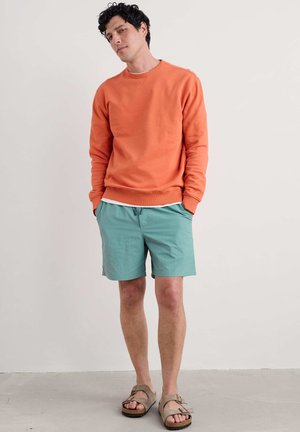 Sudadera naranja, pantalones cortos turquesa y sandalias marrones. La sudadera tiene un escote redondo y puños acanalados. Los pantalones cortos son ligeros y quedan por encima de la rodilla.