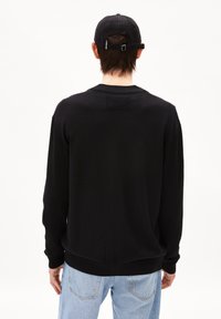 Schwarzer Pullover mit langen Ärmeln, geripptem Kragen und Bündchen, der auf der Rückseite mit einem strukturierten Patch-Detail ausgestattet ist. Getragen mit hellblauen Jeans.