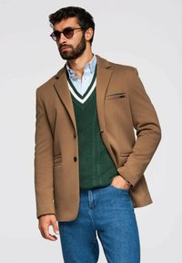 Blazer marrón sobre un suéter de punto verde con escote en V, capas sobre una camisa de color azul claro. Combinado con jeans azules y gafas de sol.