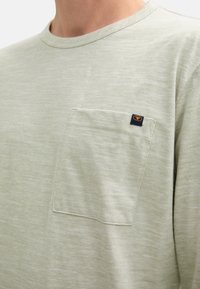 Helles grünes Langarm-T-Shirt mit strukturierter Oberfläche, rundem Ausschnitt und einer Brusttasche mit einem kleinen Logosticker.