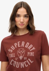 Camiseta de algodón color marrón con un estampado gráfico que presenta "SUPER DRY" y un diseño de escudo, de manga corta y con cuello redondo.