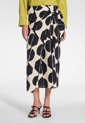 Vrouw draagt een beige wikkelrok met grote zwarte geometrische vormen, gecombineerd met zwarte sandalen met bandjes en een groen shirt met lange mouwen.