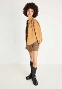 Camicia marrone con bottoni e maniche a sbuffo, abbinata a pantaloni corti a pied-de-poule e stivali di gomma neri fino al ginocchio. Il modello si trova su uno sfondo neutro.