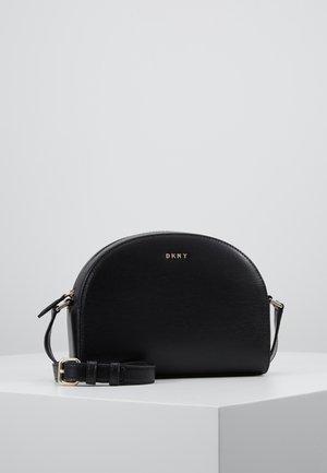 Sac bandoulière demi-lune en cuir noir avec logo DKNY doré et bandoulière réglable, présenté sur un piédestal blanc sur un fond uni.