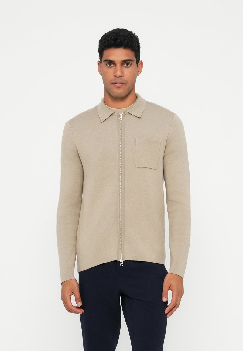 Maglione beige con zip e colletto, texture a coste, maniche lunghe e tasca sul petto. Indossato con pantaloni scuri, esposto su uno sfondo neutro.