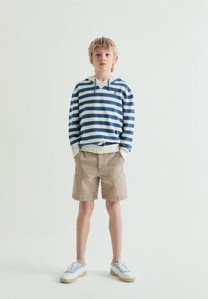 Garçon blond debout, les mains dans les poches, portant un sweat à capuche rayé bleu et blanc, un short beige et des baskets blanches sur un fond uni.