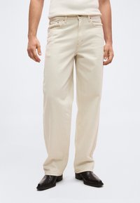 Άνδρας που φοράει φαρδιά μπεζ παντελόνια με πλαϊνές και μπροστινές τσέπες, σε συνδυασμό με σκούρα καφέ δερμάτινα παπούτσια, στέκεται μπροστά σε απλό φόντο.