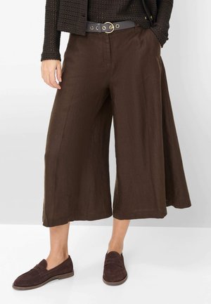 Pantaloni marroni a gamba larga e taglio corto con cintura nera, abbinati a mocassini marrone scuro e una maglia nera texturizzata.