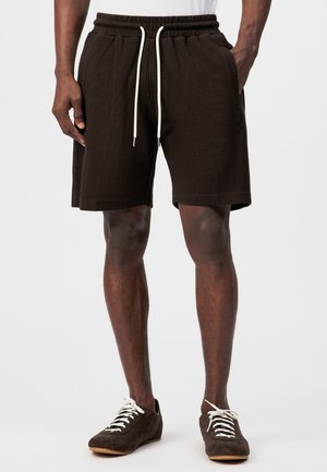 Mann trägt strukturierte schwarze Shorts mit weißen Kordeln und dunkelbraune Sneaker mit weißen Schnürsenkeln, steht vor weißem Hintergrund.