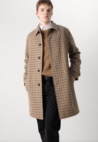 Brauner und beiger Hahnentrittmantel mit Kragen, vorderem Knopfverschluss und strukturiertem Stoff. Getragen über einem braunen Strickpullover und schwarzen Hosen.