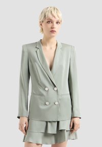 Blazer doppiopetto verde chiaro con revers, bottoni in argento e maniche lunghe, abbinato a una gonna a strati con volants.