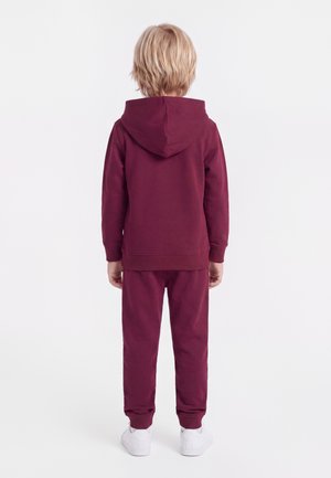 Enfant aux cheveux blonds courts, de dos, portant un sweat à capuche bordeaux et un pantalon de survêtement assorti, avec des baskets blanches sur fond blanc.