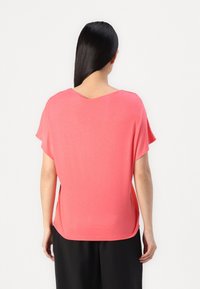 Femme aux longs cheveux noirs portant un haut ample corail à manches courtes et un pantalon noir, vue de dos sur un fond blanc.