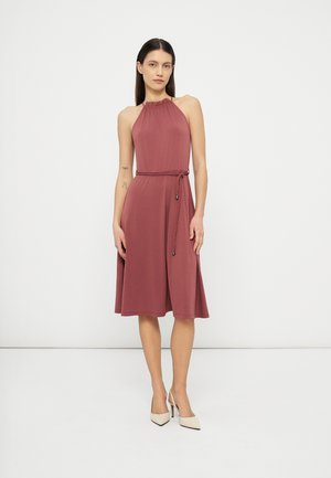 Kvinde iført ærmeløs støvet rosa knælængde kjole med flettet bælte og hvide høje hæle slingback-sko, stående mod en ensfarvet hvid baggrund.