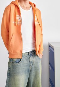 Felpa arancione con zip e testo logo bianco, abbinata a pantaloni corti in denim a gamba larga di colore blu chiaro. Maglietta bianca semplice visibile sotto.