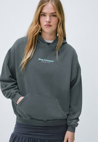 Sudadera gris oscuro de corte holgado, con bolsillo tipo canguro y texto "Stay Focused" en azul claro. Tejido suave y puños y dobladillo acanalados.
