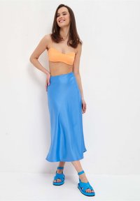 Top bandeau arancione con spalline sottili, abbinato a una gonna fluida in satin blu. La modella indossa sandali blu con lacci. Sfondo bianco.