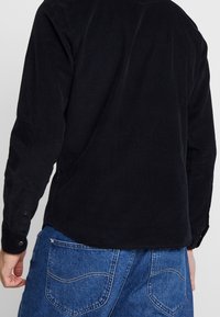 Czarna kordrowa koszula z długim rękawem, z zapięciem na guziki przy mankietach, prążkowaną fakturą i zaokrąglonym dołem, w połączeniu z niebieskimi dżinsami z tylnymi kieszeniami.