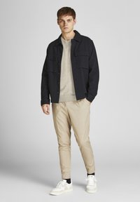 Chaqueta de camisa azul marino con bolsillos frontales, suéter gris claro, pantalones beige ajustados y zapatillas blancas con calcetines negros. Diseño simple y moderno.