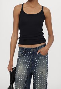 Personne portant un haut sans manches noir et un jean bleu clouté, tenant un objet en cuir noir dans la main gauche, la main droite dans la poche.