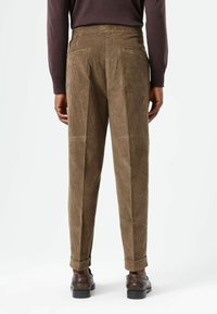 Bruine corduroy broek met rechte pijpen, voorzien van twee achterzakken en omgeslagen boorden, gecombineerd met donkere schoenen. Structuur van de stof is zichtbaar.