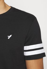 Musta lyhythihainen paita, jossa on valkoinen kolibri-logo rinnassa ja kaksi valkoista vaakasuoraa raitaa oikeassa hihassa.