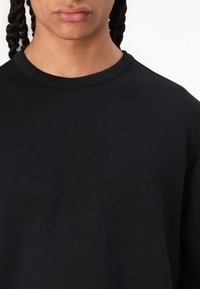 Černá mikina s kulatým výstřihem vyrobená z texturované látky, která se vyznačuje volným střihem a minimalistickým designem, podtrženým rovnými lemy a bez viditelných log.