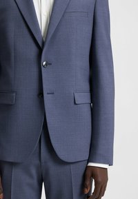 Giacca blu navy con tessuto texturizzato, chiusura con un bottone e due tasche frontali, completata da una camicia bianca sotto.