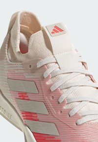 Zapatilla deportiva de color rosa claro con parte superior de punto texturizado, cordones blancos y acentos plateados, que cuenta con lazada ajustable y un parche con el logo.