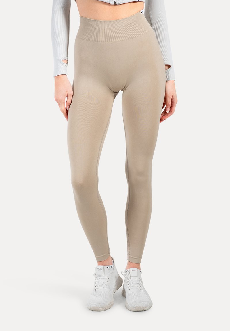 Leggings beige a vita alta, realizzati in tessuto elastico, con una texture liscia e un design aderente, abbinati a scarpe da ginnastica bianche.