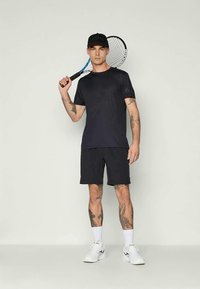 Μαύρο αθλητικό μπλουζάκι και σορτς, ορατά τα τατουάζ, κρατώντας ένα ρακέτα τένις, φορώντας μαύρο καπέλο, με άσπρα αθλητικά παπούτσια και κάλτσες μέχρι τον αστράγαλο.