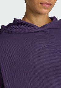 Sudadera con capucha morada de tejido texturizado; presenta un discreto logo en la parte delantera. El diseño incluye un escote amplio y ribetes acanalados.