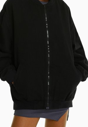 Schwarze Zip-Up-Bomberjacke mit Seitentaschen, getragen über einem kurzen dunkelgrauen Rock, Hände in den Taschen, vor weißem Hintergrund.
