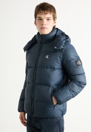 Veste matelassée bleu marine avec un col haut, fermeture éclair et logo sur la poitrine. Dispose de poches latérales et d'une capuche amovible. Texture lisse.