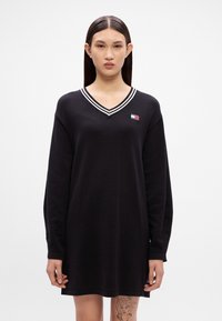 Μαύρο φόρεμα με μακριά μανίκια, V-λαιμό και λευκή ρίγα γύρω από τον λαιμό. Διαθέτει μια μικρή ετικέτα με λογότυπο στο στήθος και άνετη, χαλαρή γραμμή.