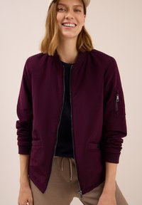 Young woman smiling wearing a beige cap, plum bomber jacket with zip and pockets, navy shirt, and beige drawstring pants.