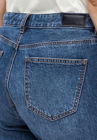 Gros plan sur la poche arrière d'un jean en denim bleu avec des coutures orange et un patch de marque en cuir noir sur la ceinture.