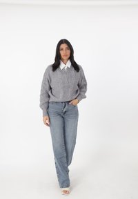 Grauer Strickpullover mit Zopfmuster, darunter ein weißes Hemd mit Kragen, kombiniert mit hoch taillierten blauen Jeans und offenen Schuhen.